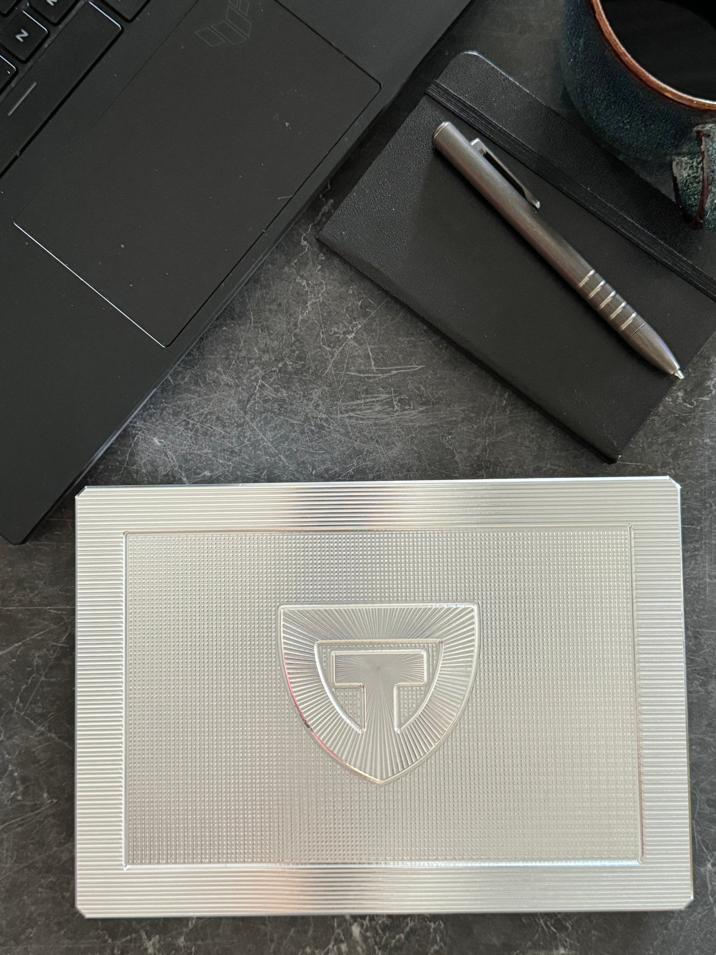 Raw machined TRT Vault Mega shown closed on a dark desk with a laptop, pen, and notebook.