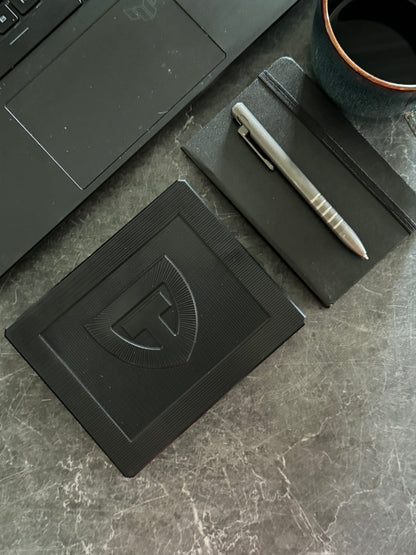 Closed anodized black Vitamin Vault Mini on a desk beside a laptop, notebook and pen.