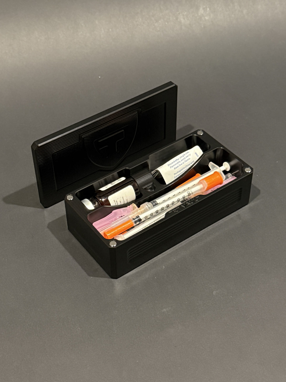 TRT Vault Micro in anodized black, shown open with vials, capped syringes and alcohol swabs.