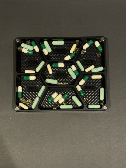 Anodized black Vitamin Vault Mini Lidless shown filled with assorted vitamins and supplements in seven labeled daily compartments.